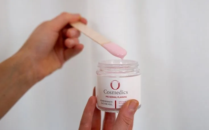 A hand holding an open O Cosmedics jar and a spatula of pink product